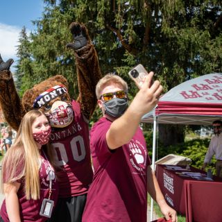 Two students wear masks and take a selfie with UM mascot Monty