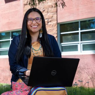 A student sits outside the Payne Family Native American Center working on her laptop. She smiles at the camera.