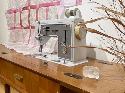 vintage sewing machine surrounded by dried grasses to the right and a paper quilt in pink and white to the left. a glass pincushion sits on the wooden table