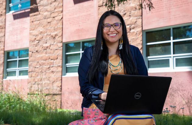 A student sits outside the Payne Family Native American Center working on her laptop. She smiles at the camera.