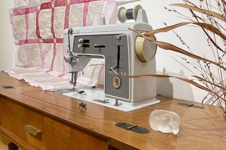vintage sewing machine surrounded by dried grasses to the right and a paper quilt in pink and white to the left. a glass pincushion sits on the wooden table