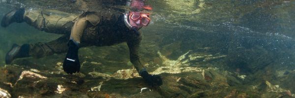 person in scuba gear with goggles and mouthpiece under the water of a creek looking at the camera