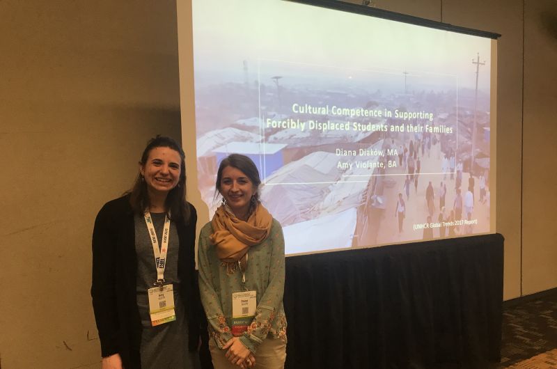 Two Graduate students standing in front of a PowerPoint entitled "Cultural Competence in Supporting Forcibly Displaced Students and Their Families""