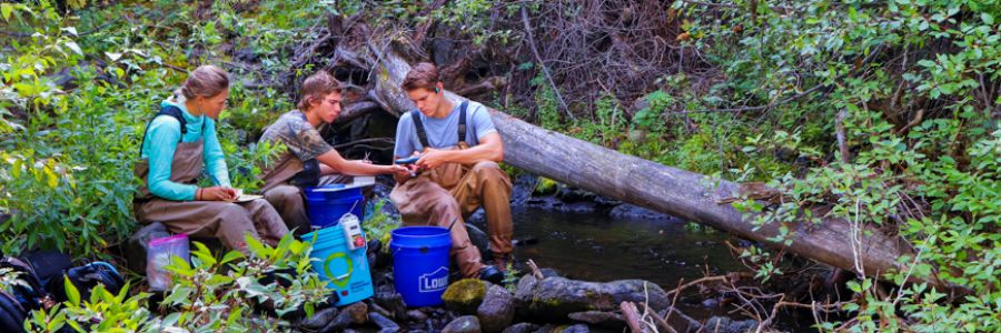 three people in waders sitting next to a creek with various scientific instruments