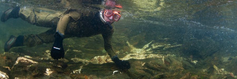 person in scuba gear with goggles and mouthpiece under the water of a creek looking at the camera