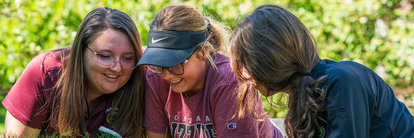 three students laying in the grass looking at a smart phone