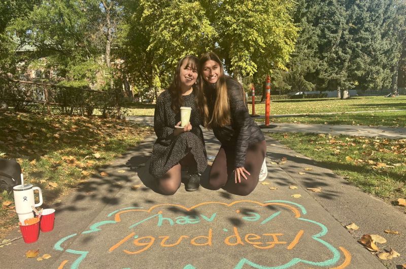 two students pose on their knees in front of a chalk phrase that is the phonetic alphabet for "have a good day"