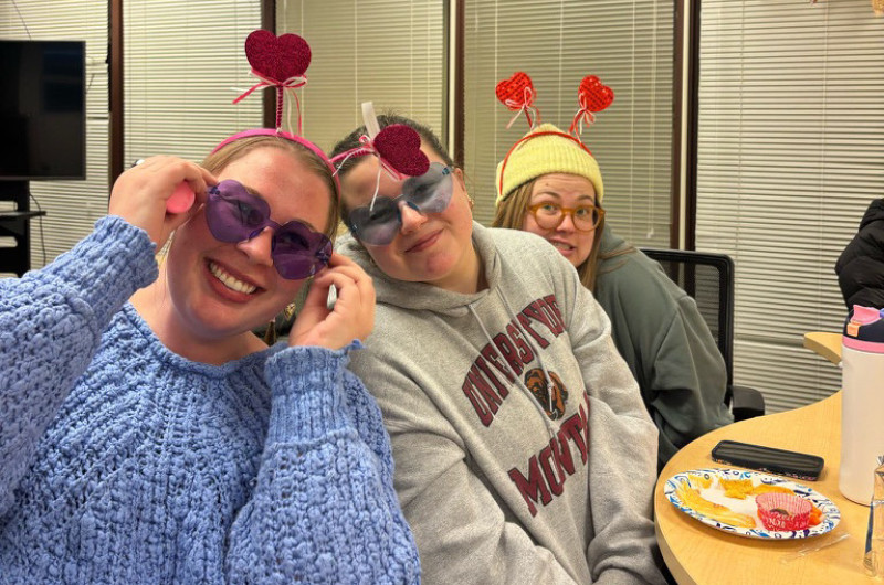 three students smile for the camera. the front and back have springy heart ear head bands. the first and second have fun blue glasses