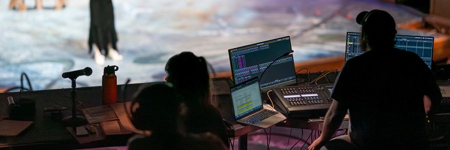 view from behind of people working on theatre audio equipment while play is happening in background