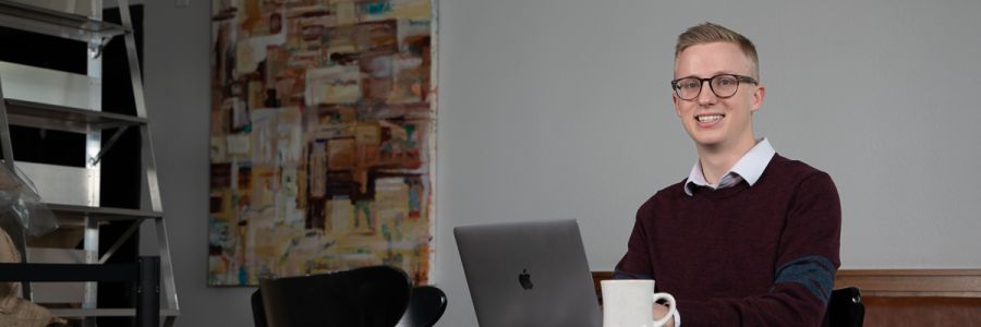 student smiling while sitting at a laptop with a cup of coffee