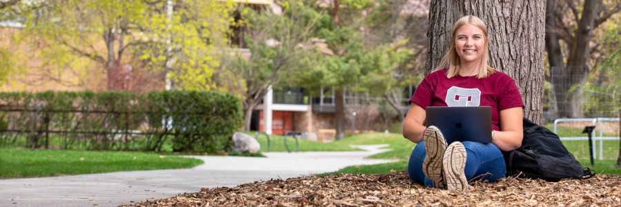 student smiling and working on a laptop outside, sitting down leaning against a tree