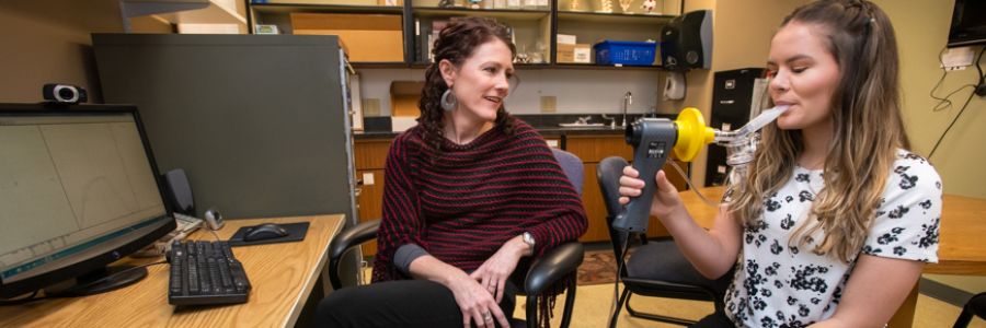 two people sitting at a computer and one blowing into a breathe/lung tester