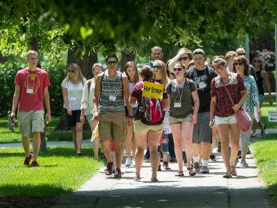A group of students walking on a sunny campus path, some holding signs and wearing name tags