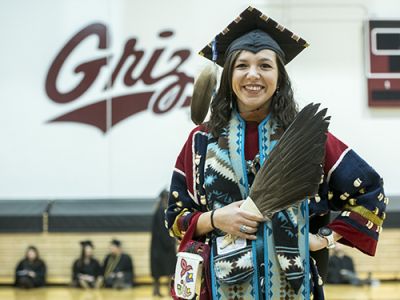Rose Bear Don’t Walk stands before the camera in her graduation regalia in front of a wall that reads: Griz