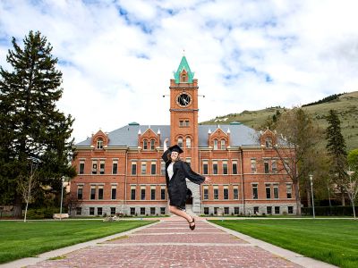 A picture of a female student jumping in front of Main Hall in her graduation outfit.