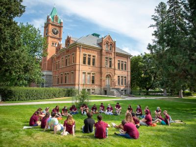 A group of students wearing maroon sits in a circle on the grass with Main Hall in the background 