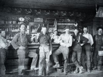 This is a very old photo of seven men standing in front of a saloon bar with bottles in the background 