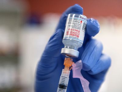 A gloved hand holds a bottle that says Moderna COVID-19 vaccine with a syringe coming out the bottom 