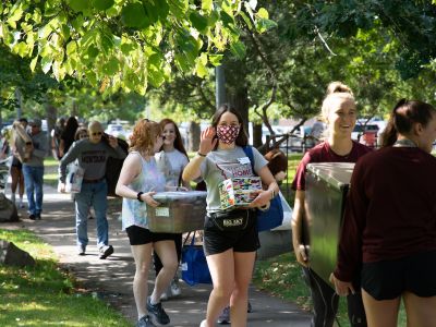 A row of students and parents carrying items walk down the sidewalk under trees