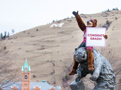 Monte holds a sign saying "Congrats Graduates" while sitting on UM's signature Grizzly Bear statue.