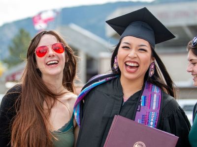 Three women smile, with one in graduation attire. 