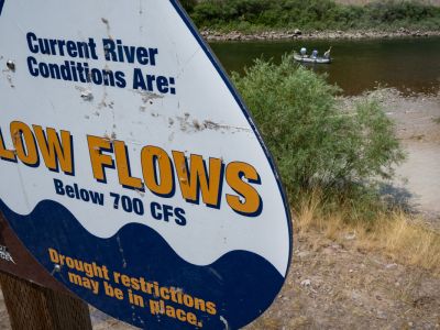 A sign near a fishing access point on the Blackfoot River warns users of low flows while a group of people fish from a raft nearby.