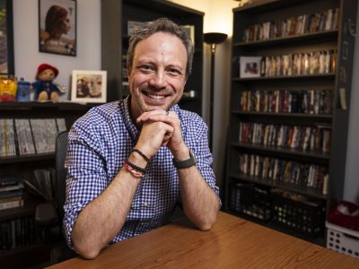91Ԫ Media Arts Adjunct Professor Tobin Addington poses for a portrait photograph in an office with a bookshelf behind him.