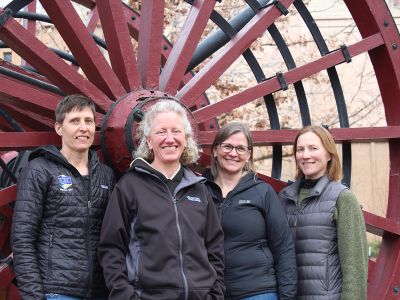 Four women stand before a high-wheel logging cart on the UM campus.