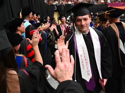 A UM student high-fives a fellow classmate at Commencement. He wears a sash that says "First-Gen."