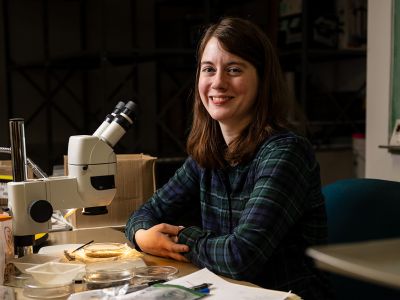 A picture of Megan Denis in a lab with a microscope.