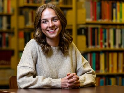 An image of UM student Lauren Schulte smiling and sitting at a desk in the library. 