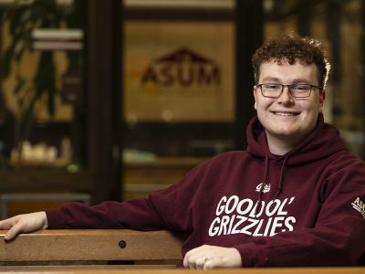 Liam Edwards sits in the University Center wearing a "Good Ol' Grizzlies" sweatshirt. The ASUM logo can be seen on a window in the background.