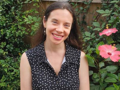 This is a headshot of Jessica Liddell, a female with long brown hair and a polka-dot shirt, standing in front of leaves and flowers 