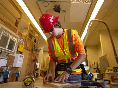 A student in an orange vest and hard hat measures a piece of wood at a construction site 