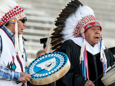 Two Native Americans, in eagle feather headdresses, sing and carry drums.