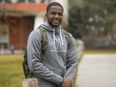 This is a portrait of a young Black man in a grey sweatshirt and a camo backpack smiling and a blurred out building in the background 