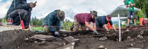 three people kneeling and digging at an artifact site while two others watch