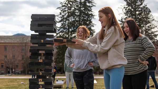 Students gathered around a large jenga tower with one student pulling a jenga piece. 