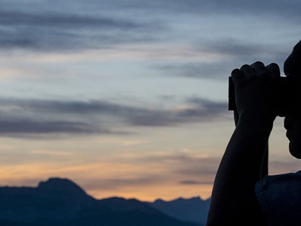 A silhouetted hunter looks through binoculars as the sunrise illuminates the sky behind him. 