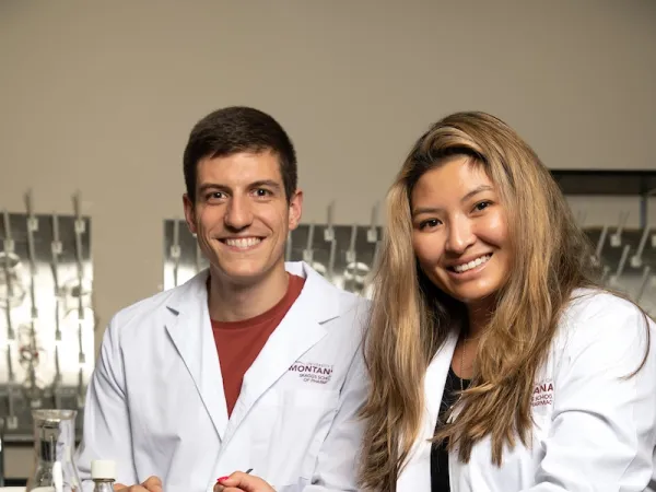 Two UM Pharmacy students in white lab coats seated at a laboratory bench with glassware and a medication bottle.