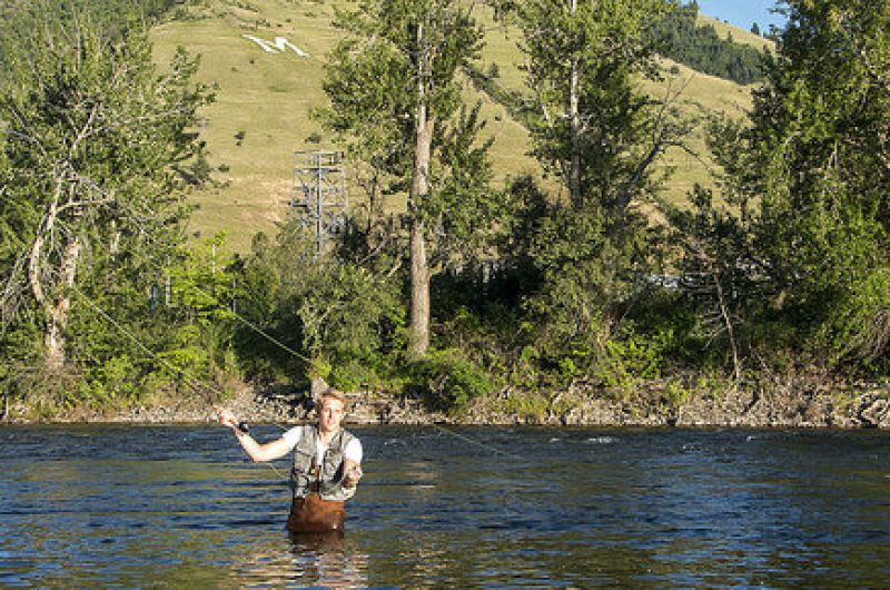 Person fly-fishing while wading in the Clark Fork in Missoula, with trees and the M on the hillside in the background.