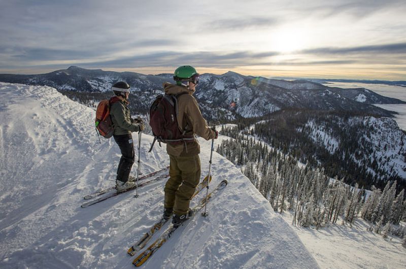 Two skiers pause on a snowy ridge at Snowbowl, looking out over a mountain valley at sunset