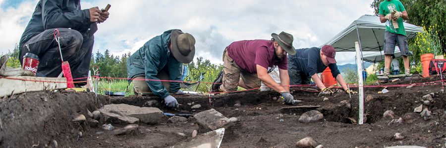 three people kneeling and digging at an artifact site while two others watch