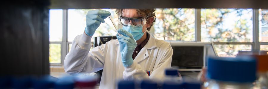 person with googles, mask and other protective equipment working on a sample in a test tube