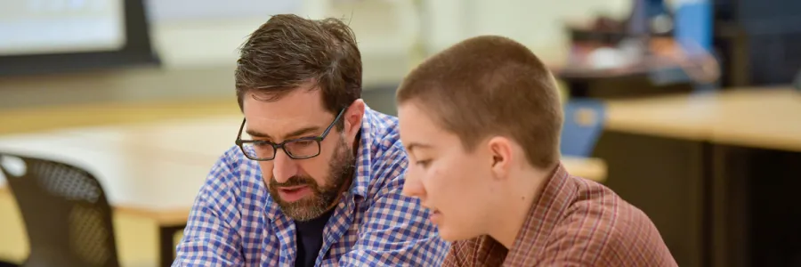 A ؿ student and professor wearing collared, button-up, long sleeve shirts sit at a table having a discussion.