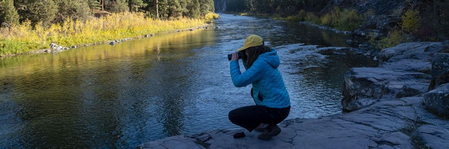 person knelling down and taking a picture on the rocky bank of a river